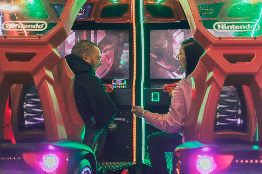 Two people sitting on arcade motorcycle rides facing each other in front of a gaming screen in an arcade, surrounded by colorful neon lights.