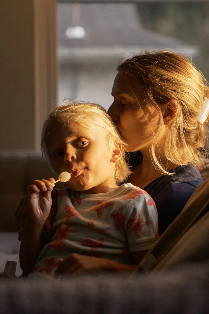 A woman kissing a young boy on the cheek while he holds a lollipop in his mouth, sitting on a couch near a window with natural light.