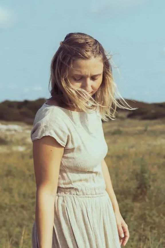 A young woman in a beige dress standing in a field with a clear blue sky.