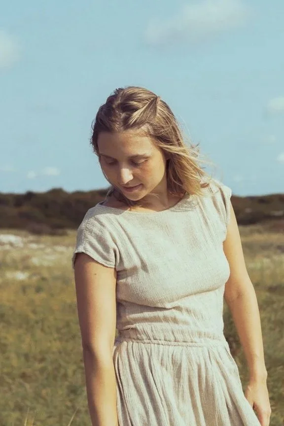 A woman in a light-colored dress standing outdoors in a grassy field under a partly cloudy sky.