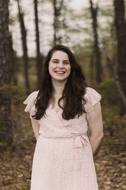 A young woman with long dark hair and a light pink dress standing outdoors in a wooded area, smiling at the camera.
