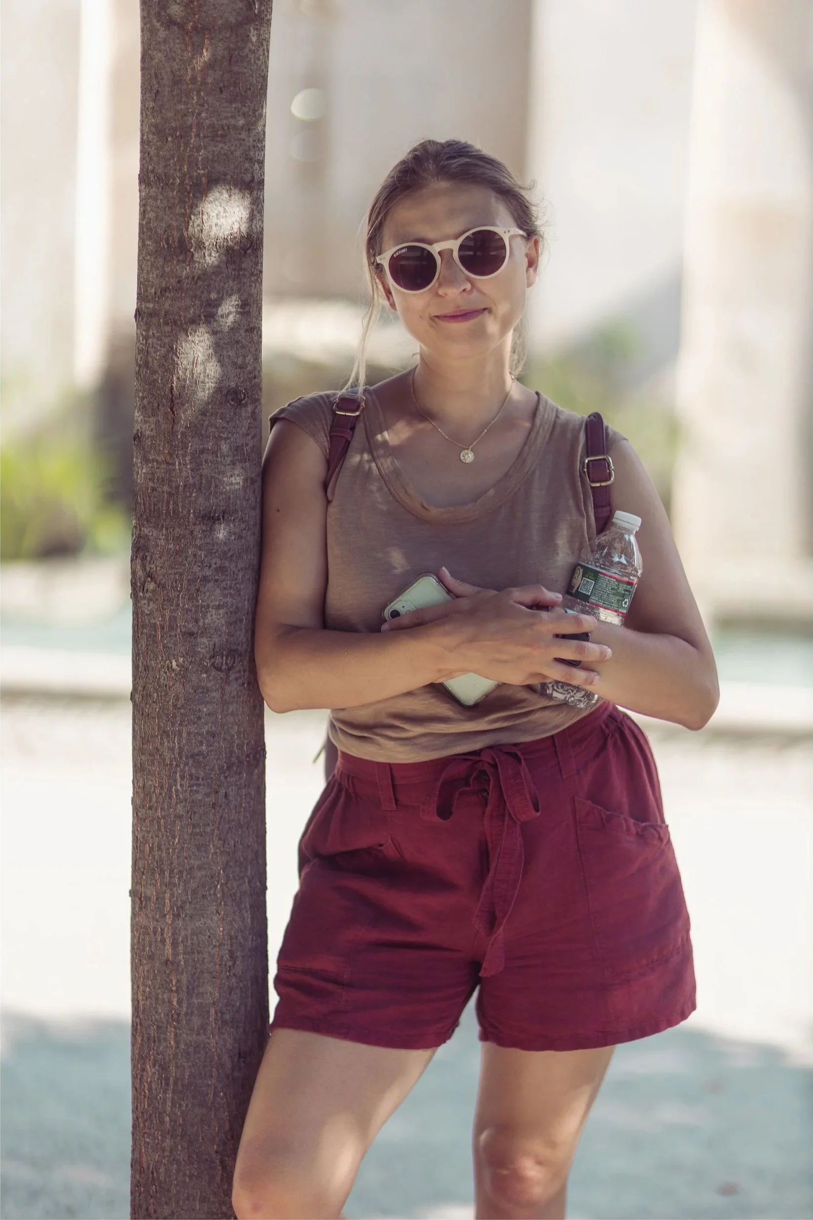 A woman standing outdoors between a tree and a blurred background. She is wearing sunglasses, a brown sleeveless top, maroon shorts, and holding a water bottle and smartphone.
