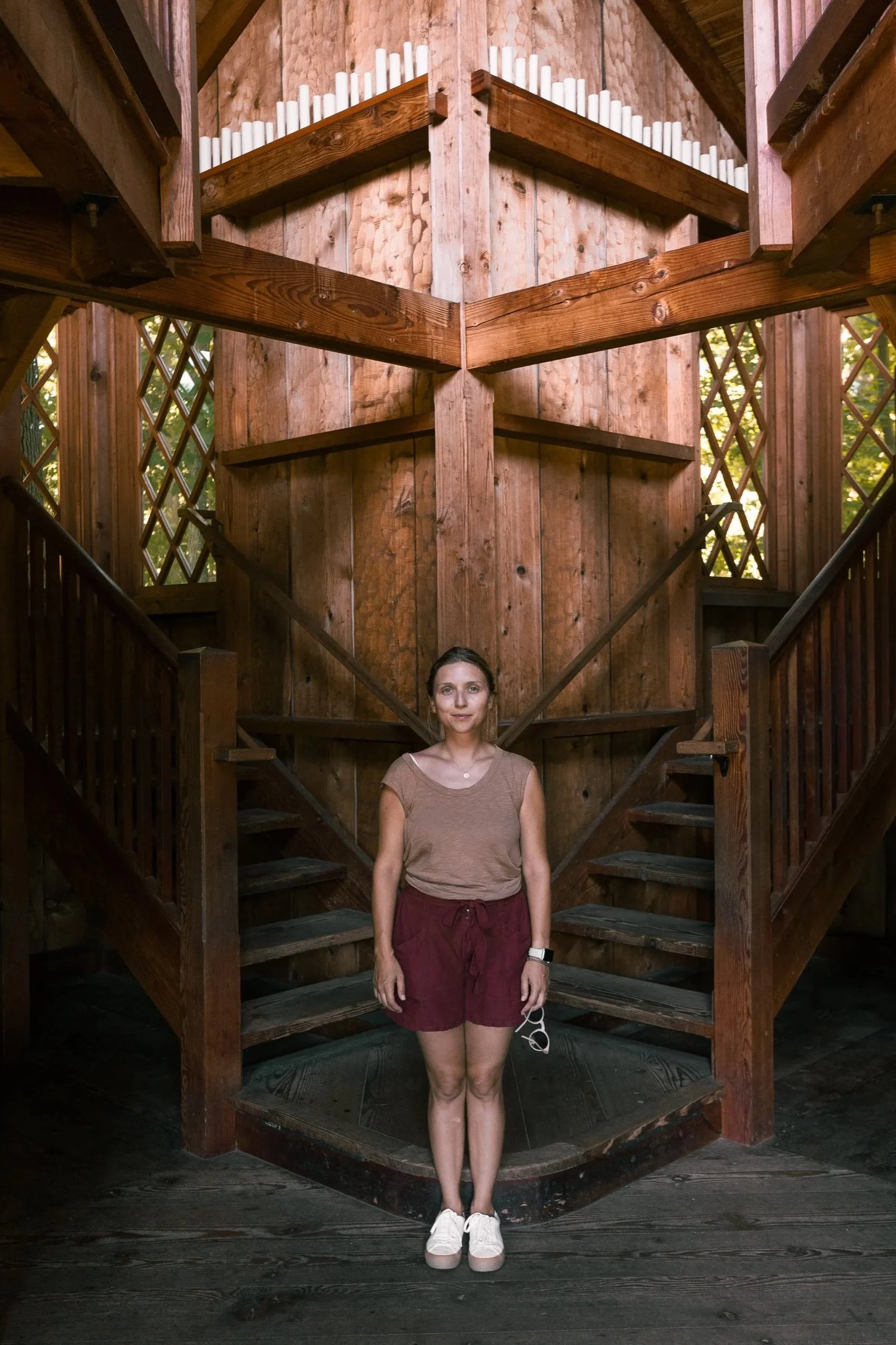 A woman standing on wooden stairs inside a rustic wooden structure with a large wooden paneled wall behind her. The structure has decorative lattice windows on both sides and a triangular roof. The woman is wearing a brown t-shirt, maroon shorts, and