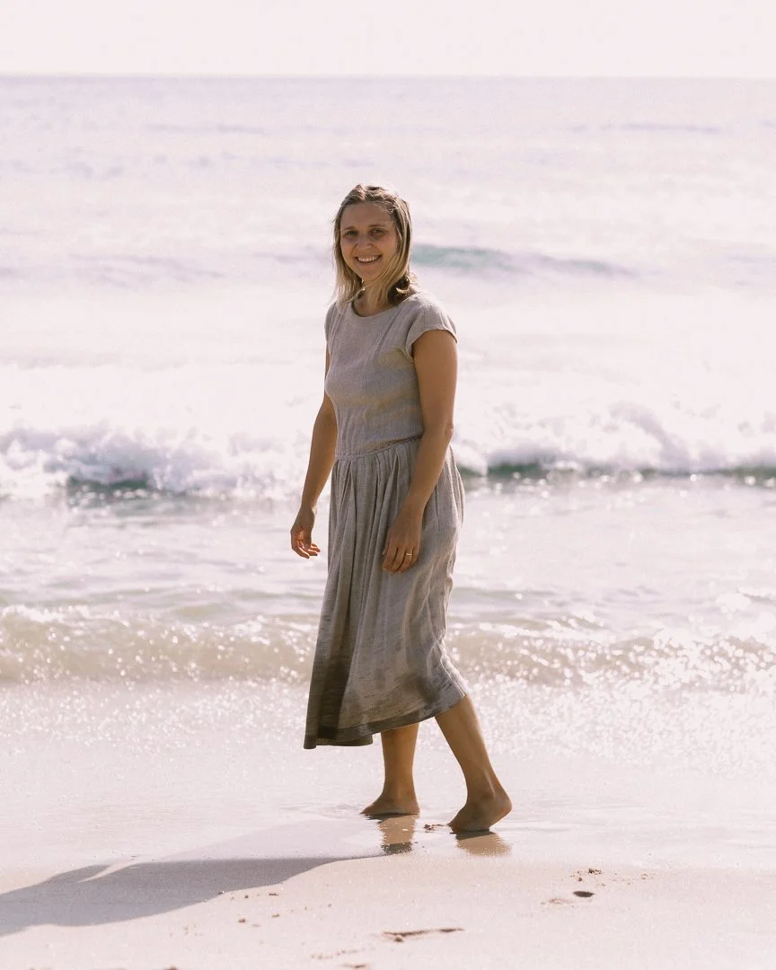 A woman in a beige dress walking barefoot on the beach near the ocean, smiling at the camera.