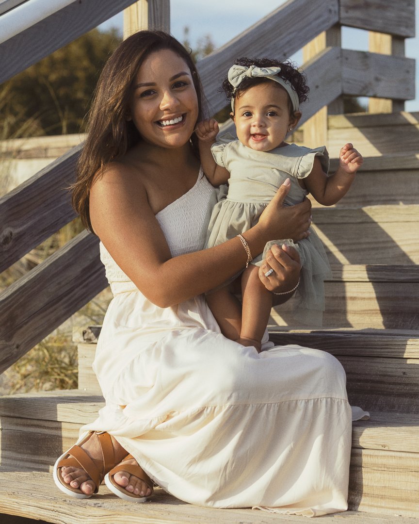 A woman sitting on wooden stairs outside, holding a smiling toddler girl with curly hair and a headband, both dressed in light-colored clothing, during daytime.