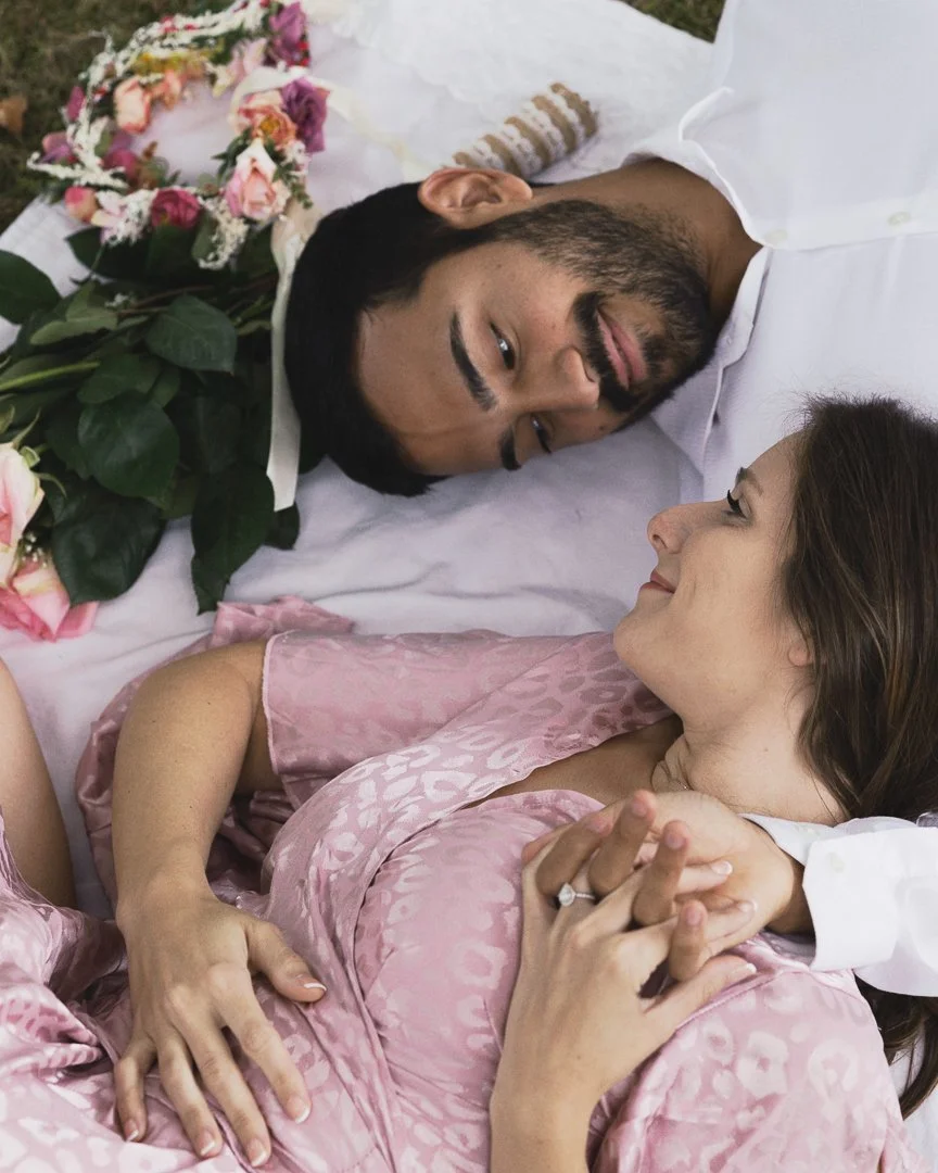 A man and woman lying together on a bed, gazing at each other, with the woman smiling and holding hands, and a bouquet of flowers nearby.