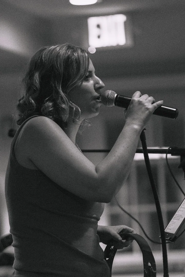 A woman singing into a microphone at an indoor event, with an exit sign in the background.