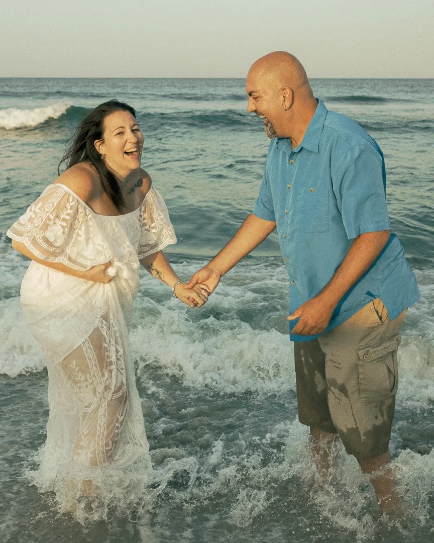A couple holding hands and laughing in the shallow ocean water at the beach during sunset.