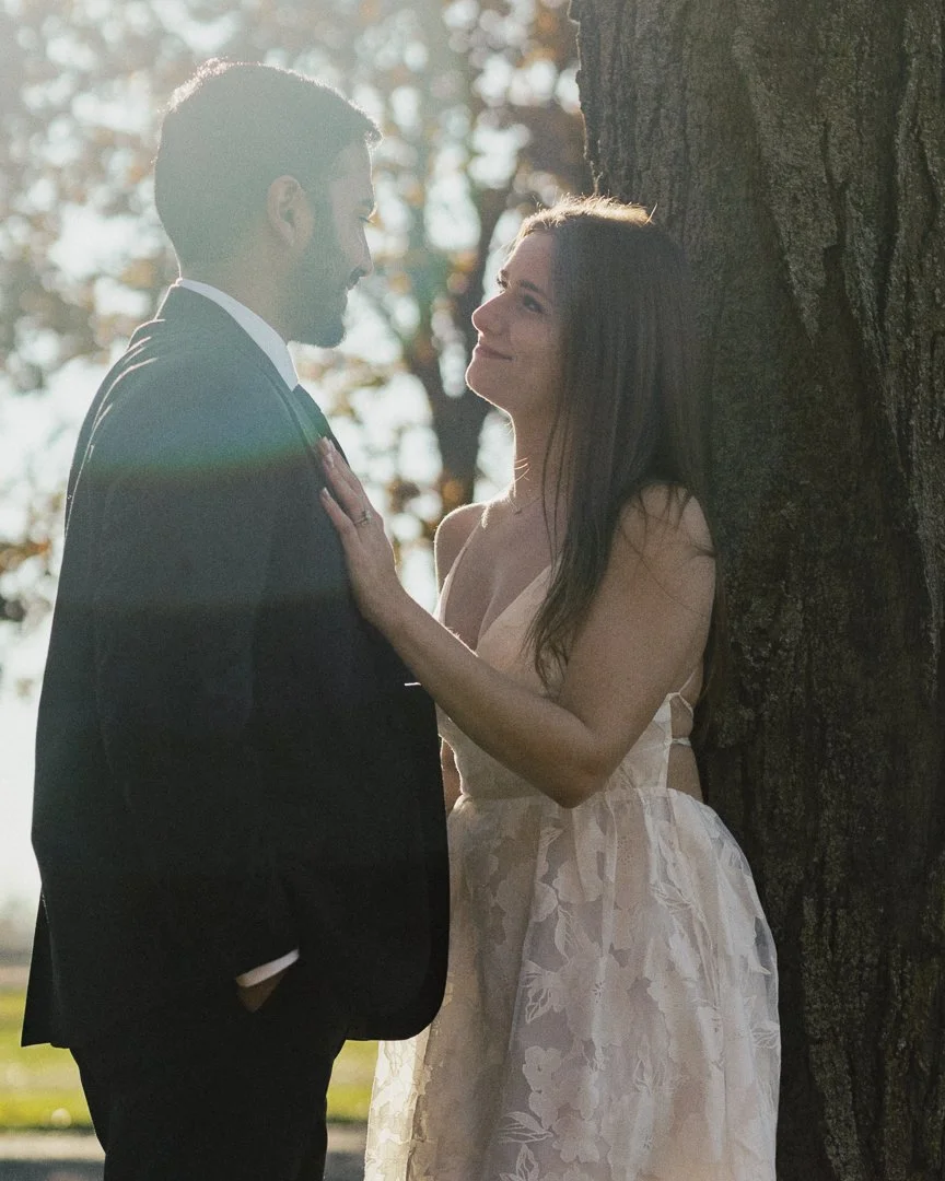 A man in a suit and woman in a dress standing close to each other outdoors near a tree, smiling and gazing into each other's eyes.