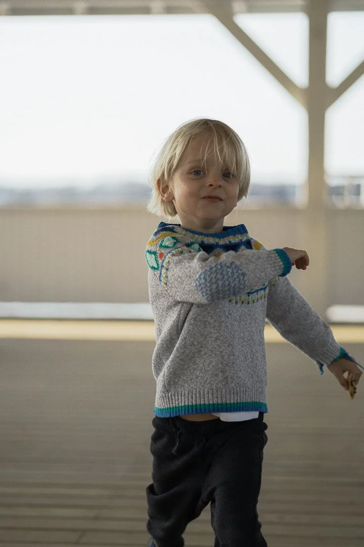 A young boy with blond hair wearing a knit sweater with colorful patterns, dark pants, standing on a deck with a blurred background.