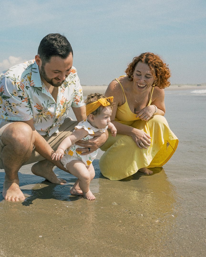 A family of three at the beach, including a father, mother, and baby girl, enjoying and playing with the water.