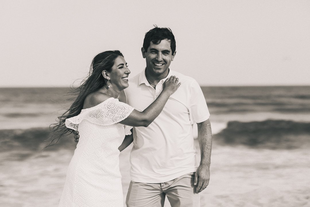 A black and white photo of a smiling couple on the beach. The woman, wearing a white dress, is laughing and has her arm around the man's shoulder. The man, wearing a white shirt, is smiling at the camera.