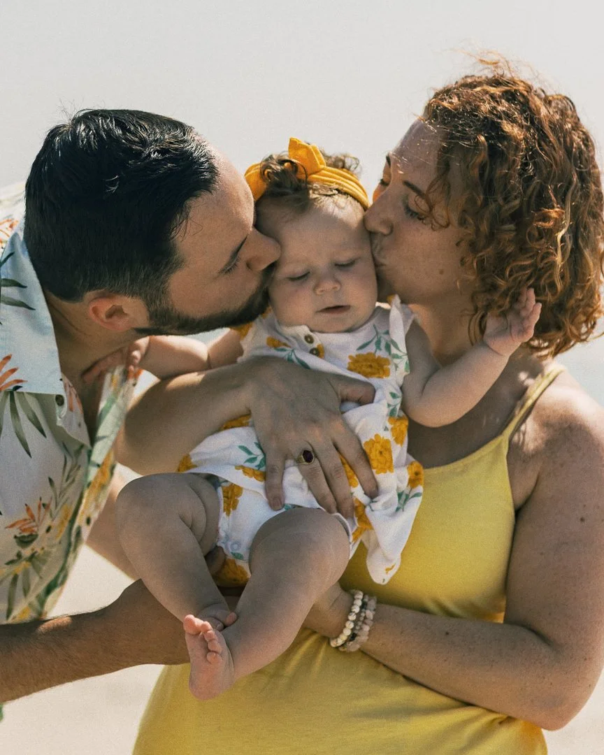 A family of three on the beach, with two adults kissing a smiling baby girl on each cheek.