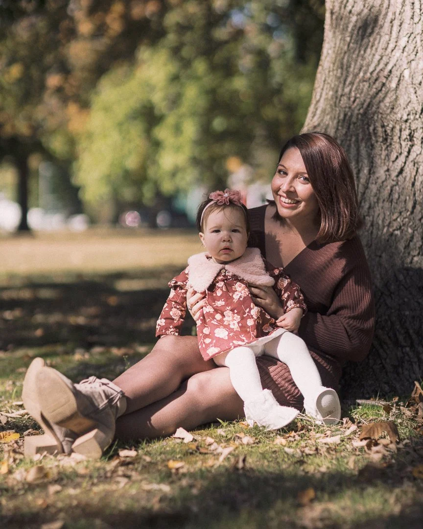A woman and a baby girl sitting on grass outdoors next to a tree, smiling, with autumn leaves scattered around.