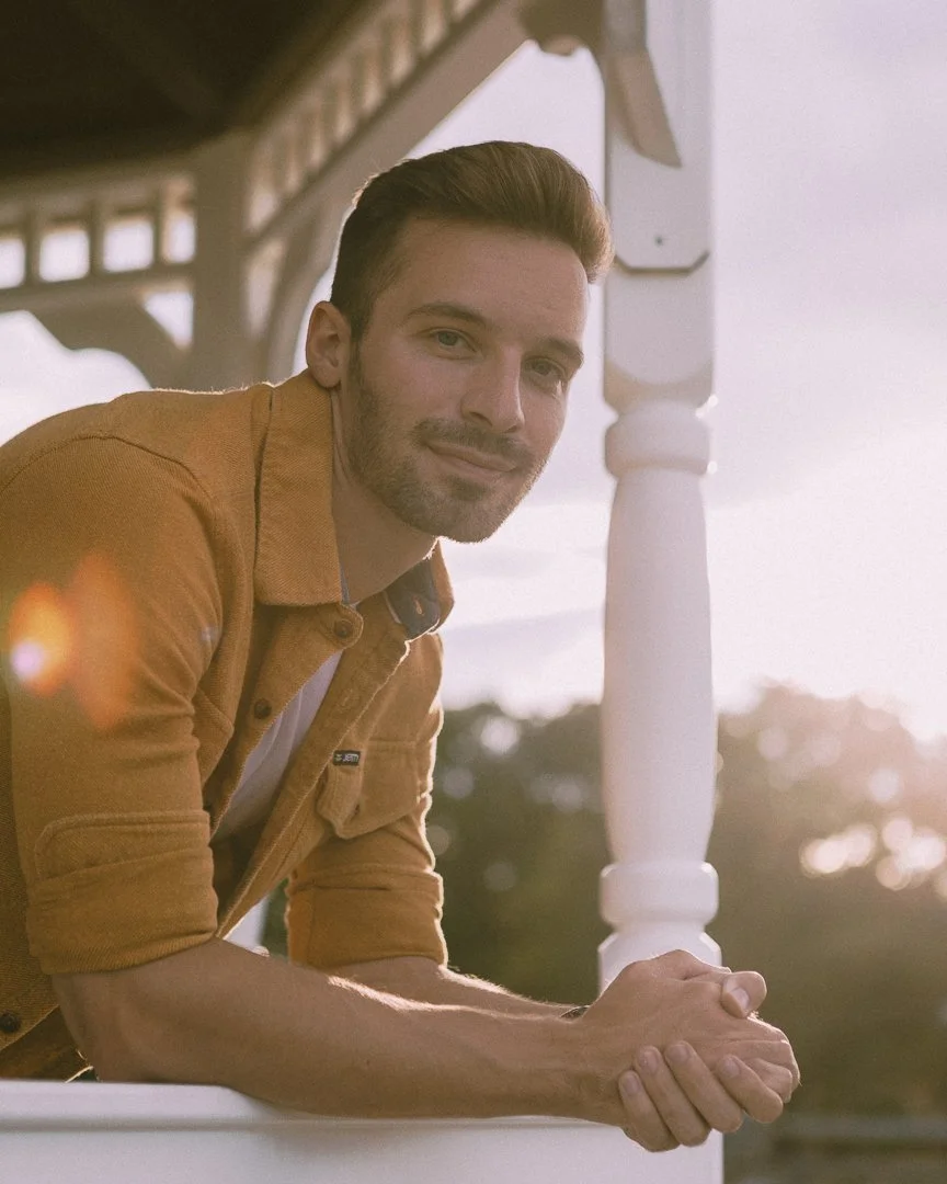 A man with brown hair and a beard leaning on a porch railing at sunset, wearing a tan jacket and smiling at the camera.