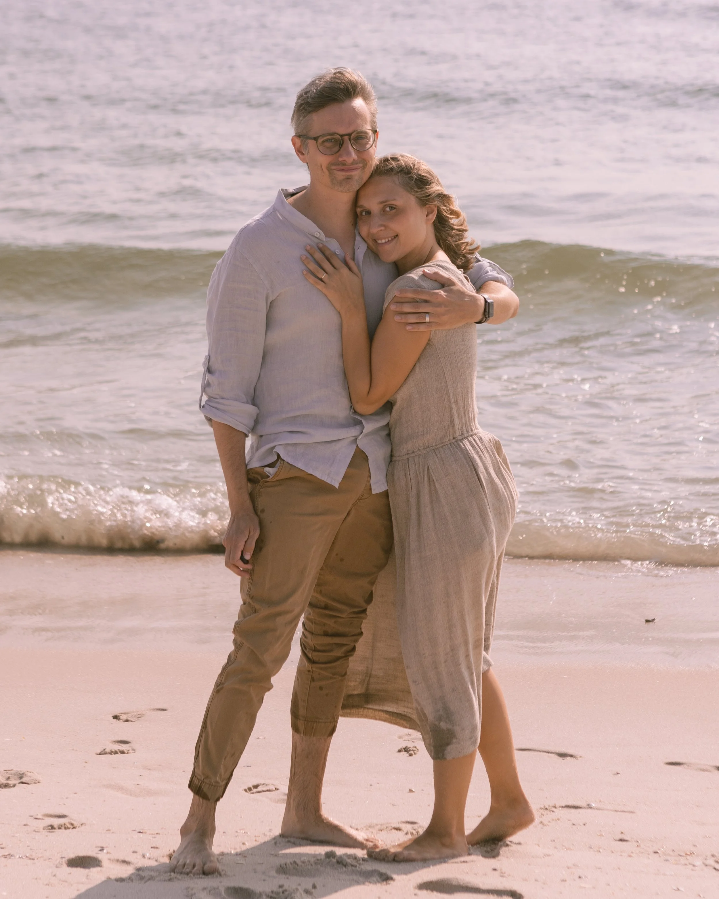 A couple hugging on a sandy beach with ocean waves in the background, smiling at the camera.