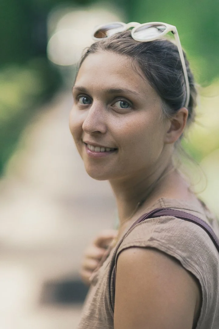 A smiling woman with light skin, blue eyes, and brown hair pulled back, wearing sunglasses on her head and a tan shirt, outdoors with blurred green foliage in the background.