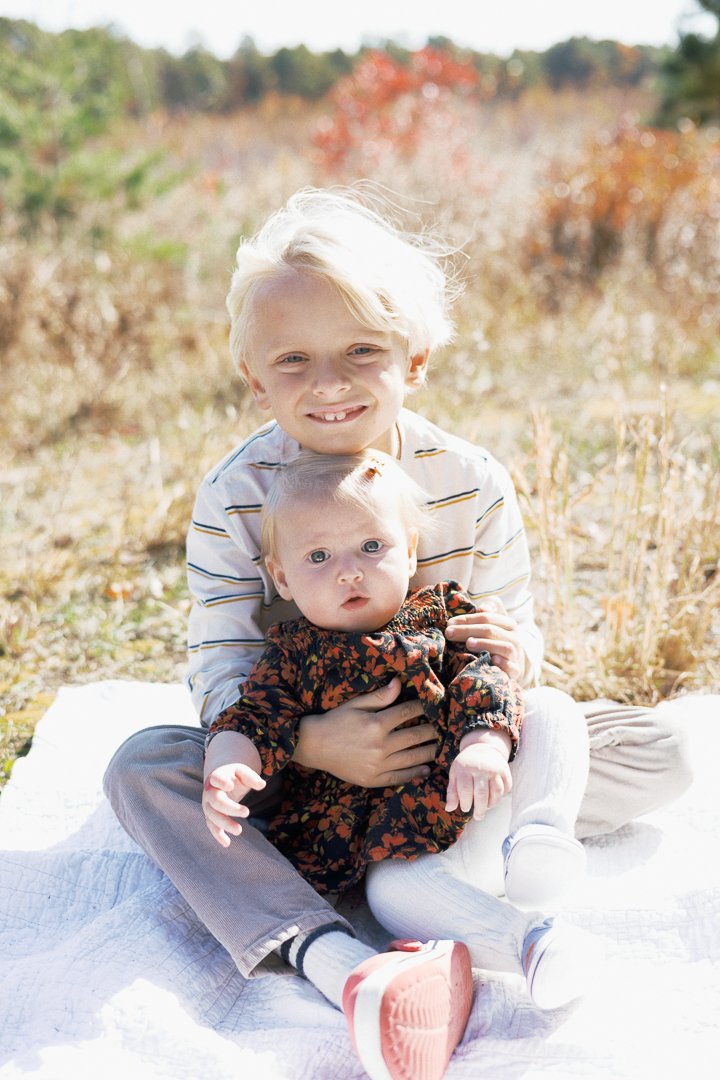 Two young children sitting on a white blanket outdoors in a field during fall, with one older boy smiling and holding a younger girl.
