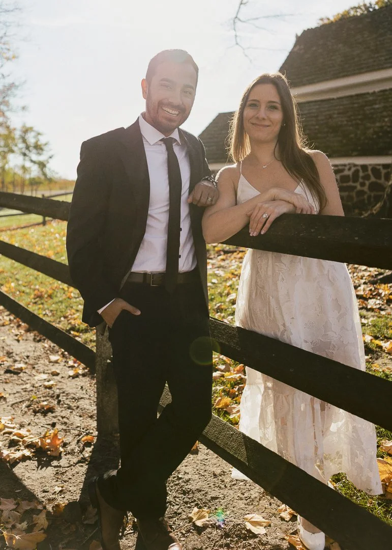 A man in a suit and a woman in a white dress stand outdoors near a wooden fence on a fall day, smiling at the camera.