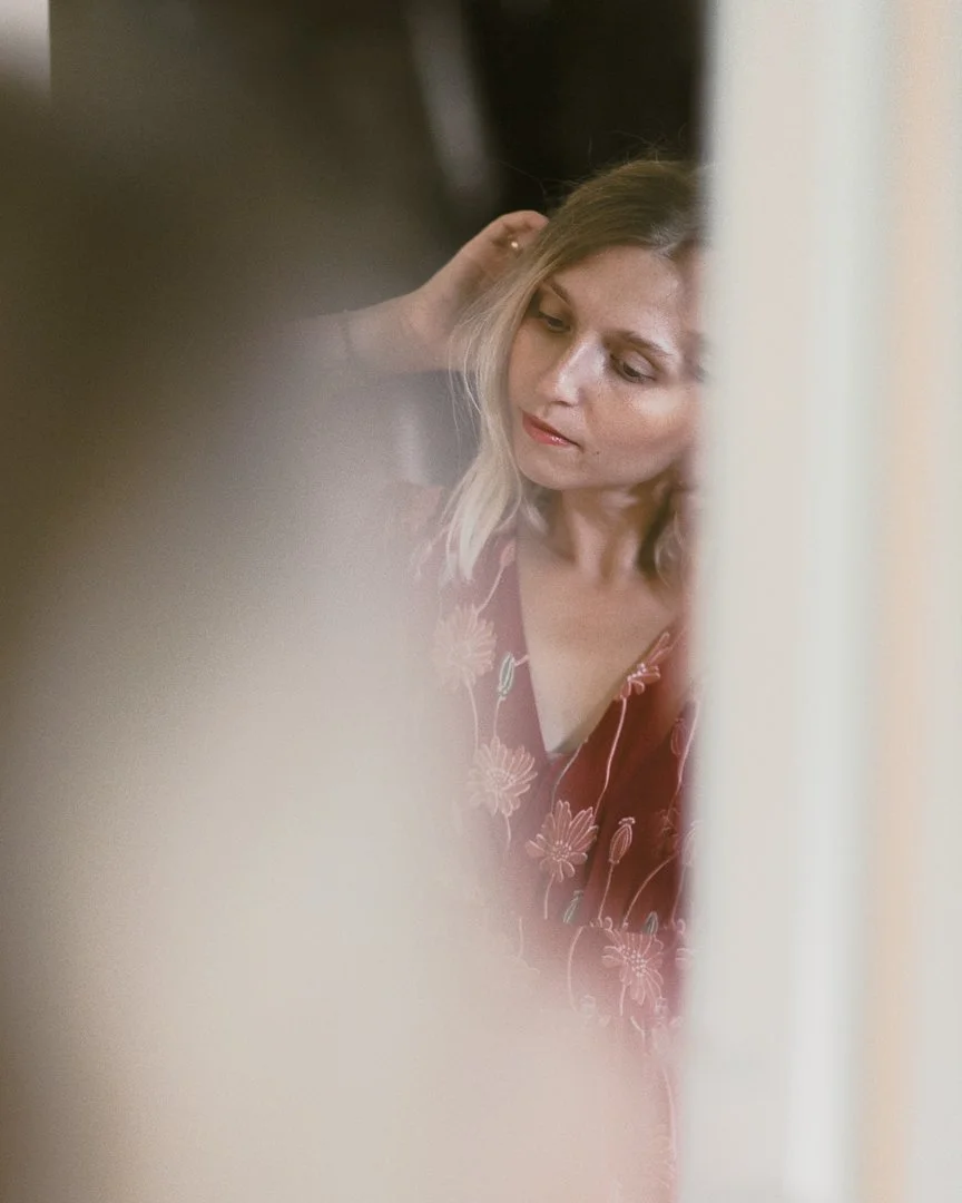 A woman with blonde hair looking down while touching her hair, seen through an obscured or blurred foreground.