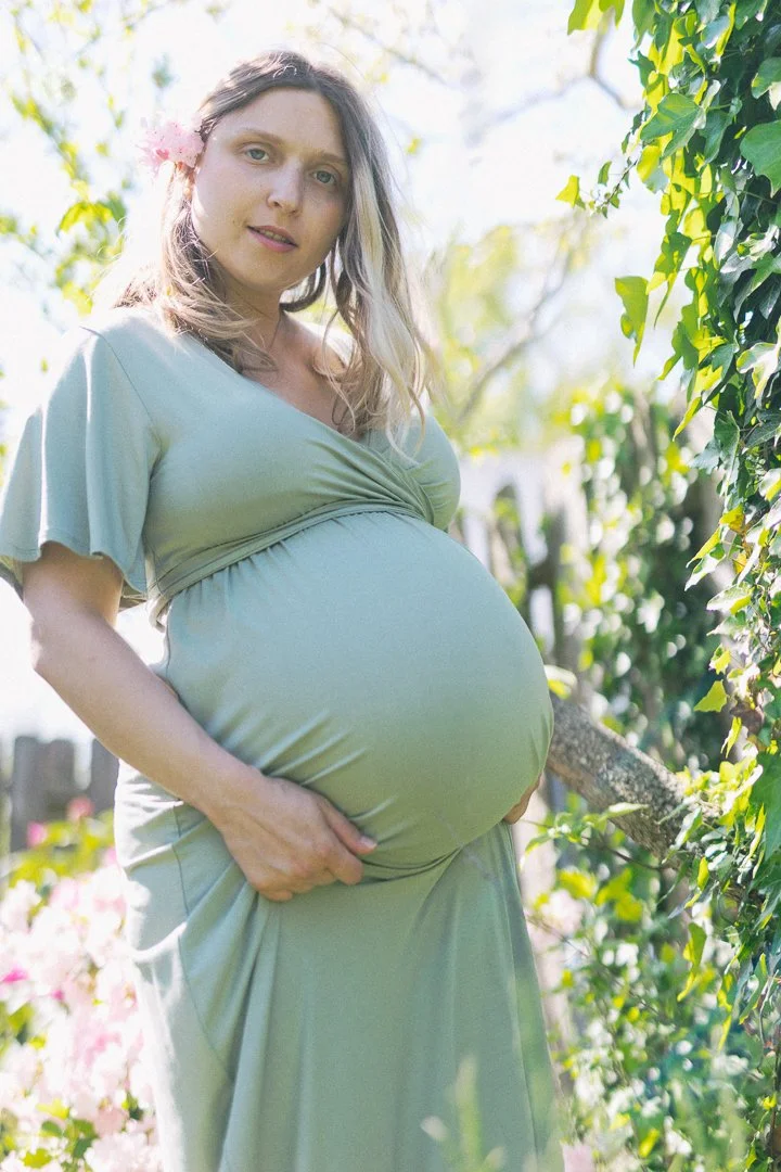 A pregnant woman holding her belly outdoors surrounded by greenery and flowers, wearing a light-colored dress, with a flower in her hair.