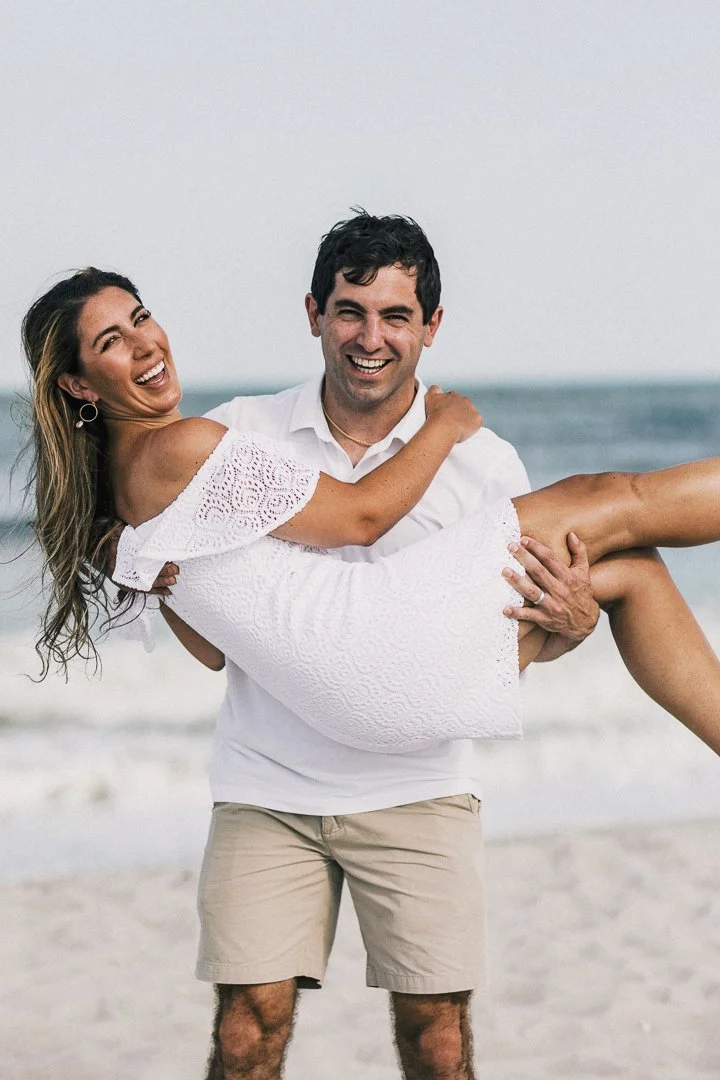 Man carrying woman in white dress on a beach, both smiling.