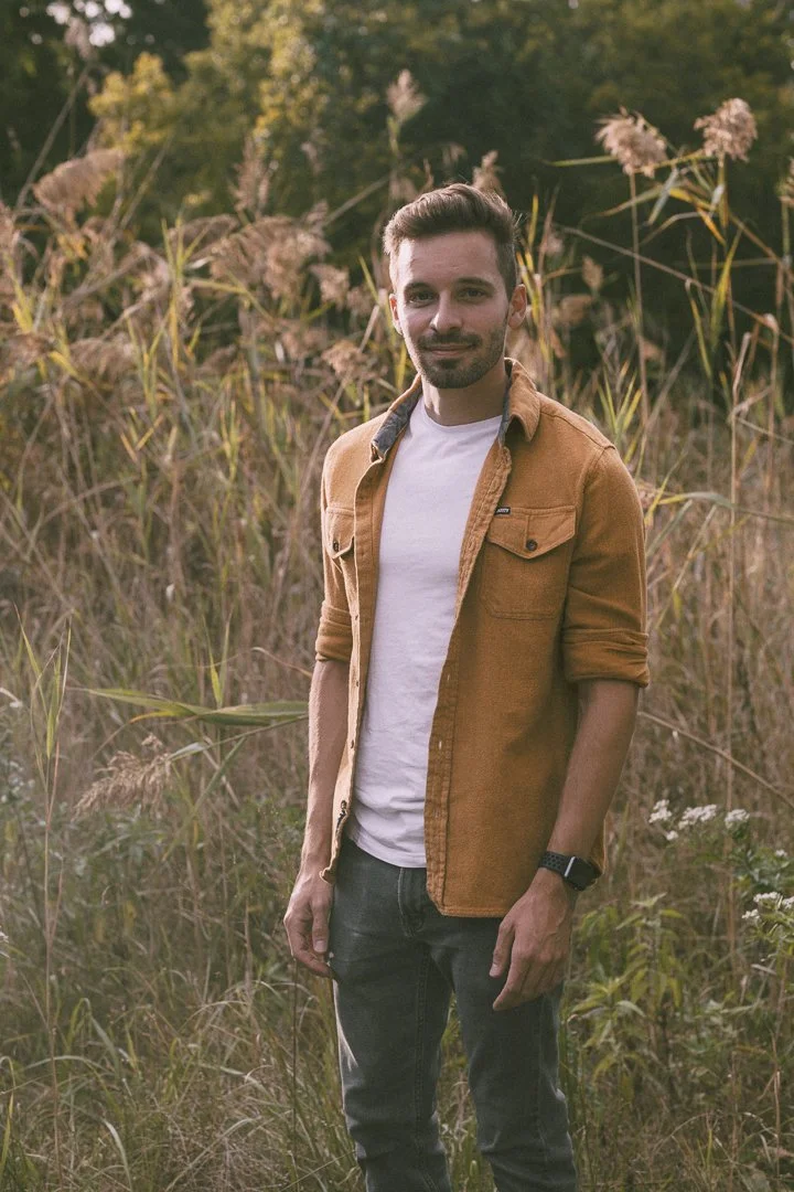 A young man with short brown hair and facial hair standing outdoors among tall grasses and wild plants, wearing a white t-shirt, an orange-brown jacket, and a wristwatch, during sunset or late afternoon.