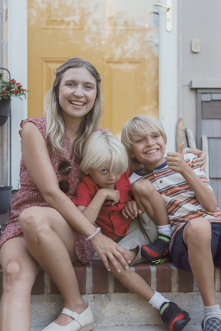 A smiling woman sitting on steps with two young boys, all relaxed and happy outside a house with a yellow door.