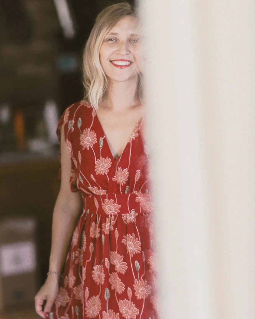 A smiling woman in a red floral dress standing behind a white curtain, indoors.