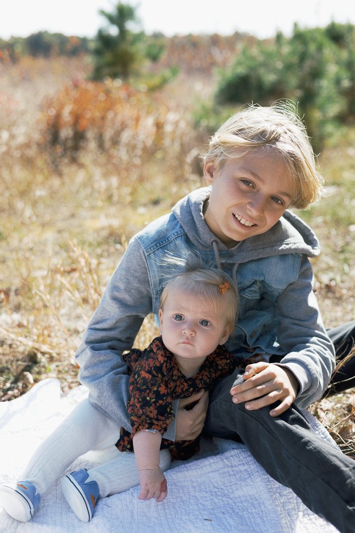 Two children, an older boy and a younger girl, sitting outdoors on a white blanket in a natural setting with autumn foliage in the background. The boy is smiling and holding the girl, who has a serious expression.