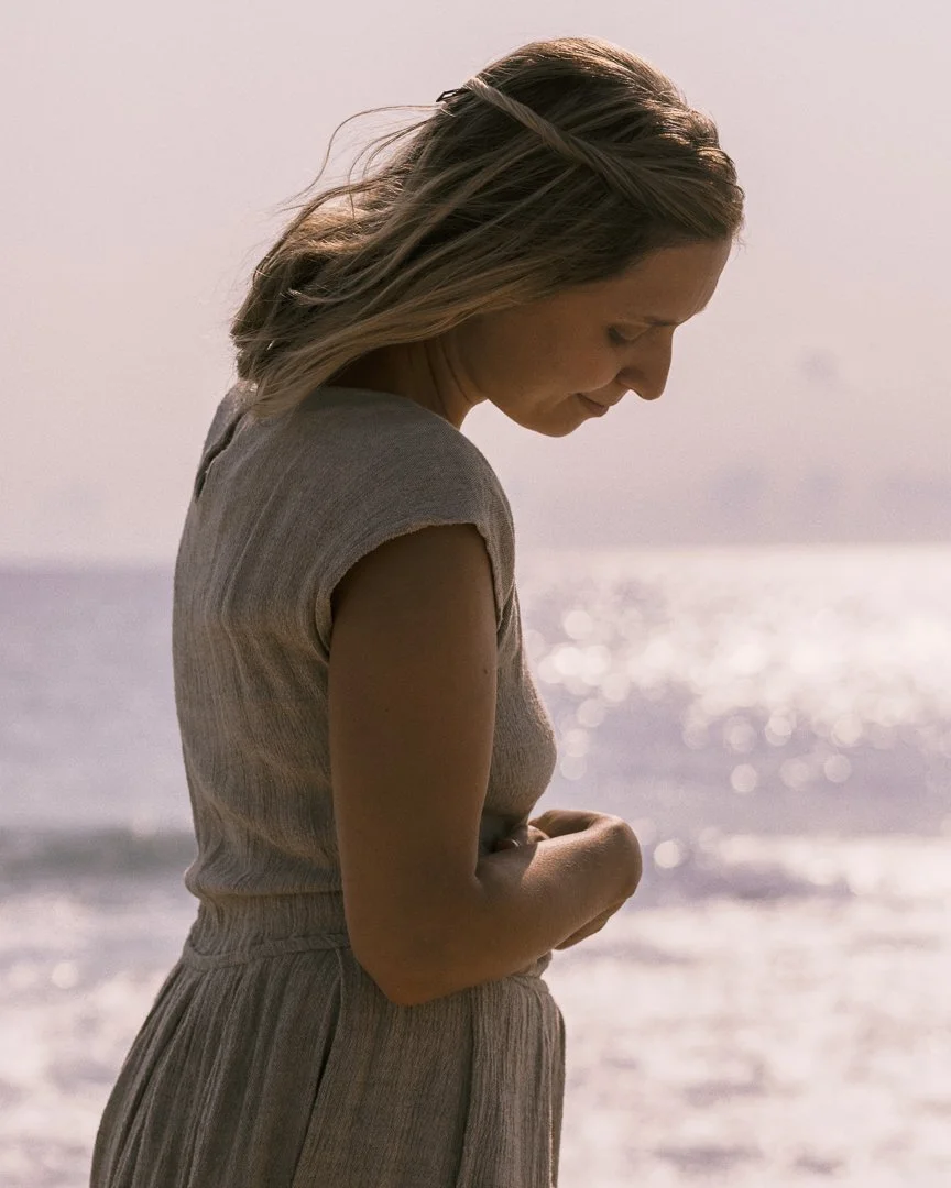 A woman with blonde hair standing by the ocean, looking down with a gentle expression.