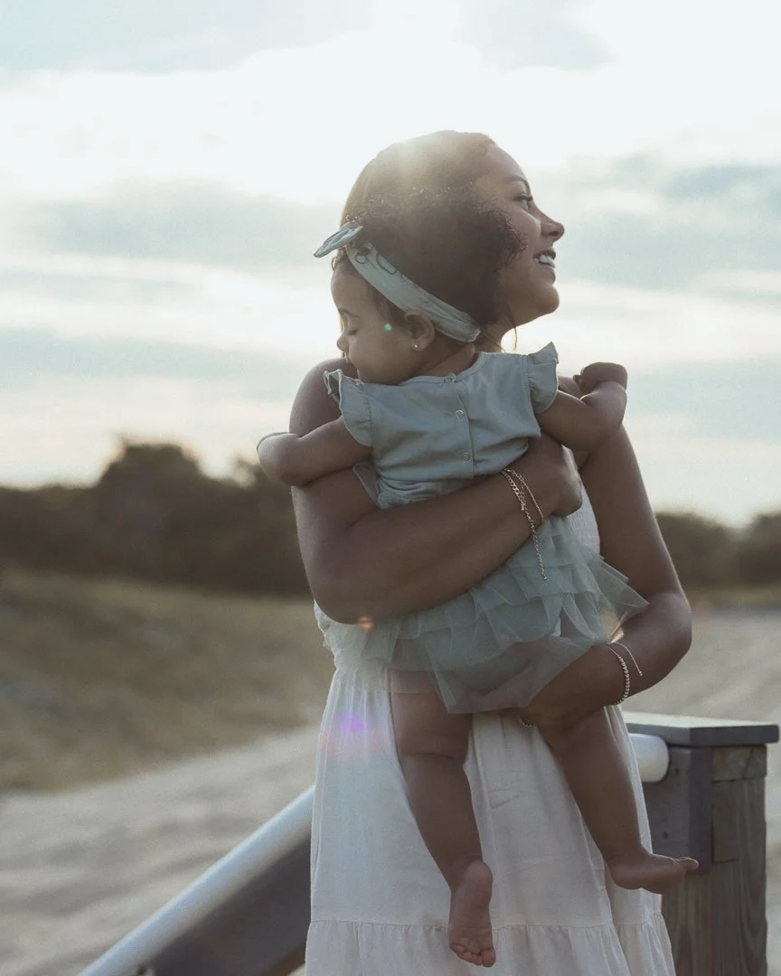 A woman holding a young girl in her arms outdoors, both smiling, with a cloudy sky and landscape in the background.