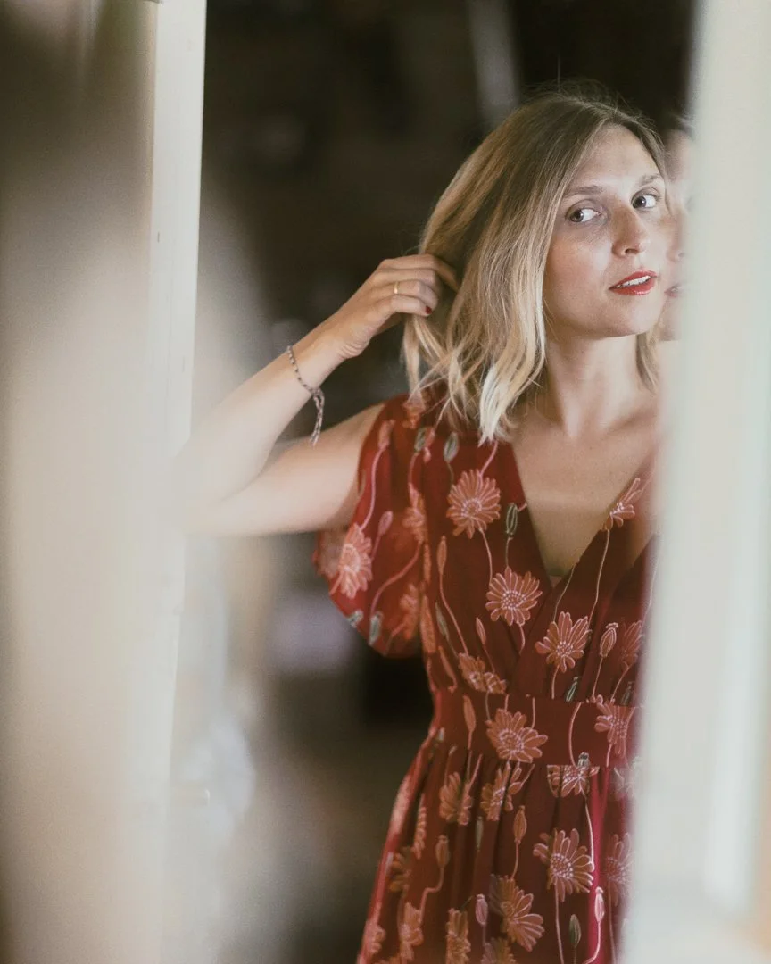 A woman with shoulder-length blonde hair and red lipstick is looking in a mirror while adjusting her hair. She is wearing a red dress with a floral pattern.