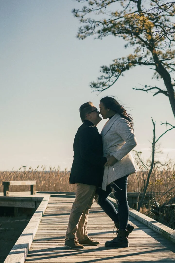 A couple kissing on a wooden dock outdoors, surrounded by trees and tall grass, during sunset or late afternoon.