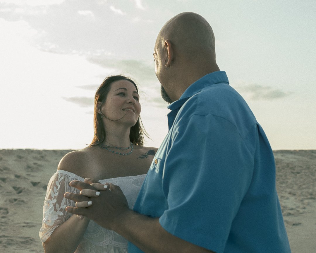 A couple dancing on the beach during sunset, with the woman smiling and looking at her partner.