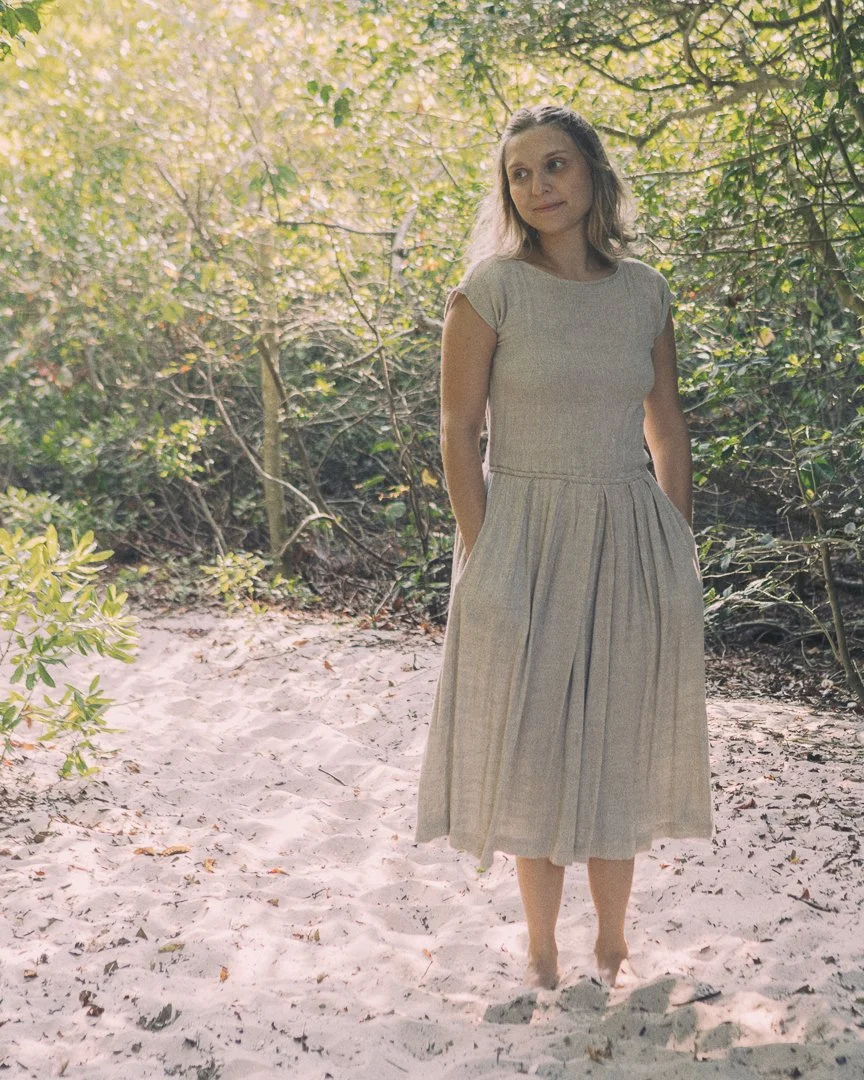 A woman with light brown hair in a grey dress standing barefoot on sandy ground with green trees and foliage in the background.