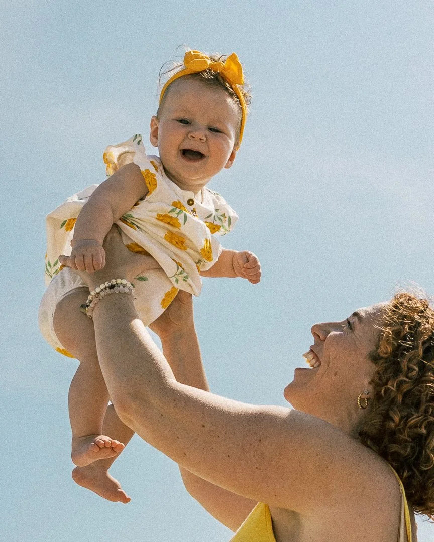 A woman with curly hair holding a smiling baby girl in the air against a clear blue sky. The baby is wearing a yellow headband and a white dress with yellow flowers.