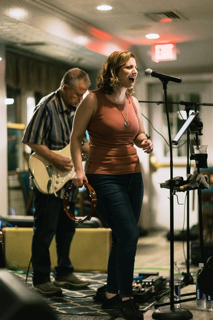 A woman singing passionately into a microphone at a live music performance, holding a tambourine, with a guitarist playing behind her in an indoor venue.