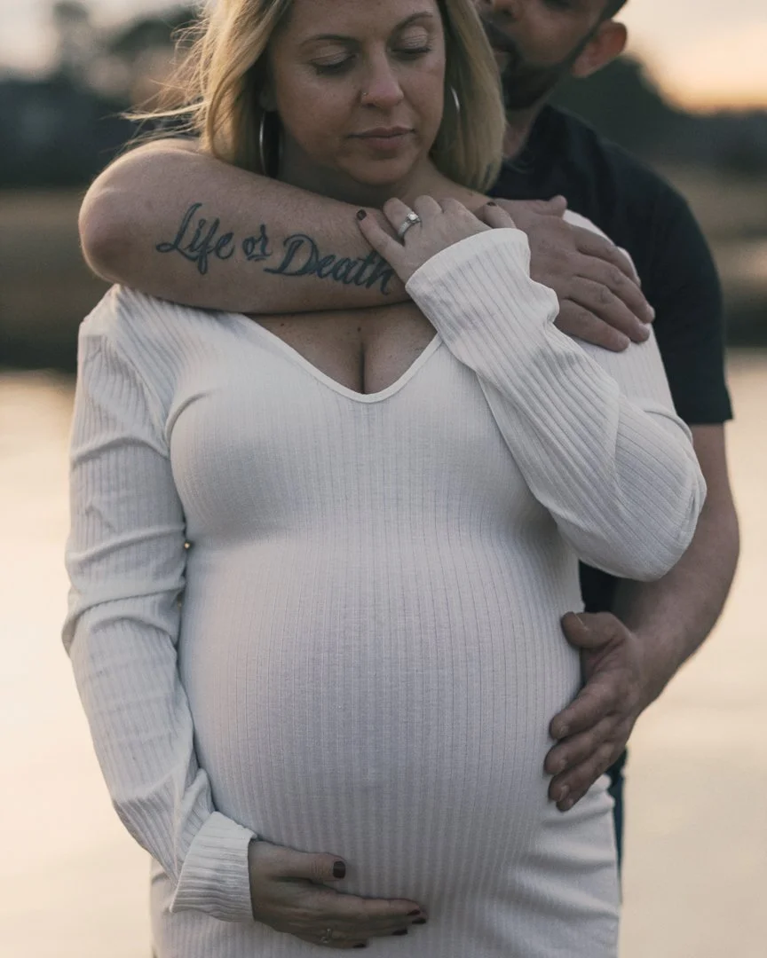 A pregnant woman with blonde hair, wearing a white ribbed dress, is being embraced by a man from behind, both by a body of water at sunset. The woman has her eyes closed, and a tattoo on his arm reads 'Life or Death.'