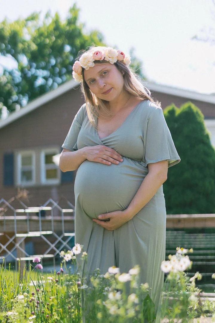 Pregnant woman with a flower crown on her head standing in a garden, holding her belly, with a house and trees in the background.