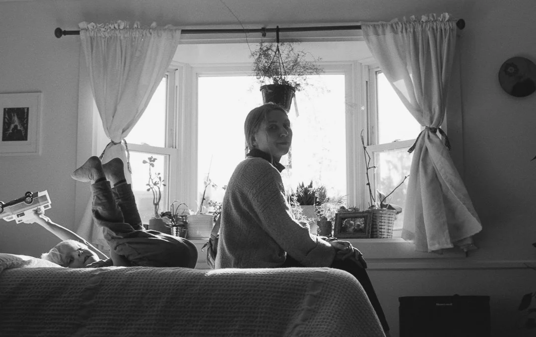A woman sitting on a bed in front of a window with white curtains, with a child lying on the bed with legs up in the air. The window has various potted plants and framed photos on the windowsill, and a hanging plant is visible above. The room appears