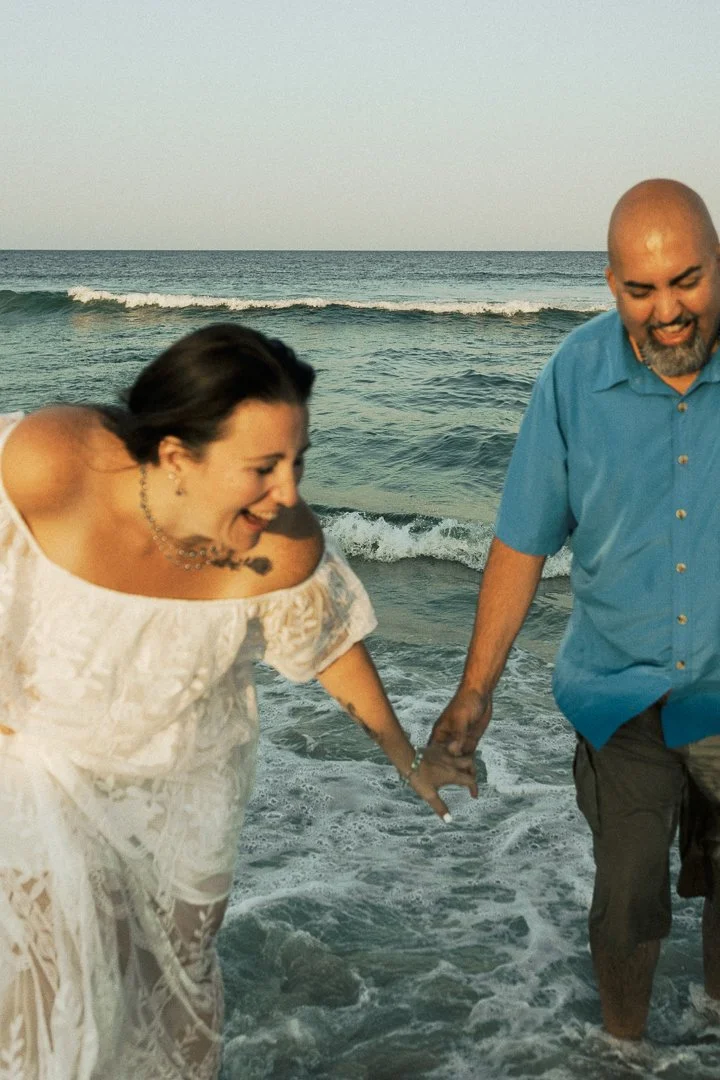A smiling couple holding hands at the beach, walking in the water with the ocean and sky in the background.