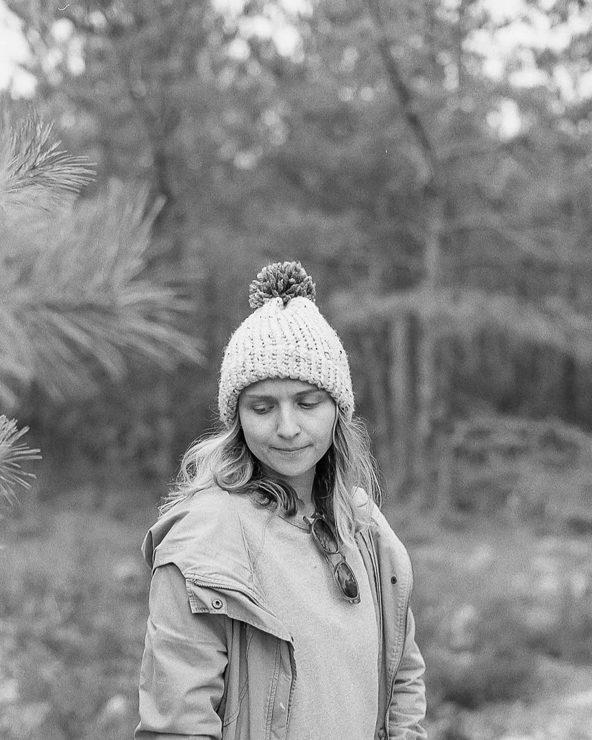 A woman in a knit beanie with a pom-pom stands outdoors near pine trees in a forest setting.