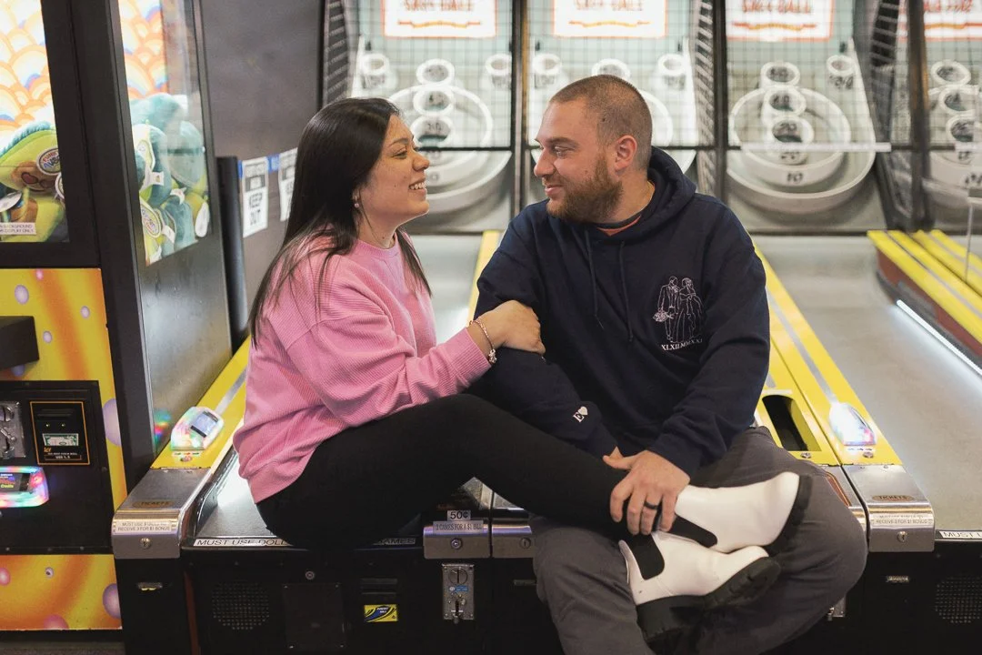 A woman and man sitting on an arcade game machine in an arcade, smiling at each other, with the woman sitting with her legs draped over the man's lap.