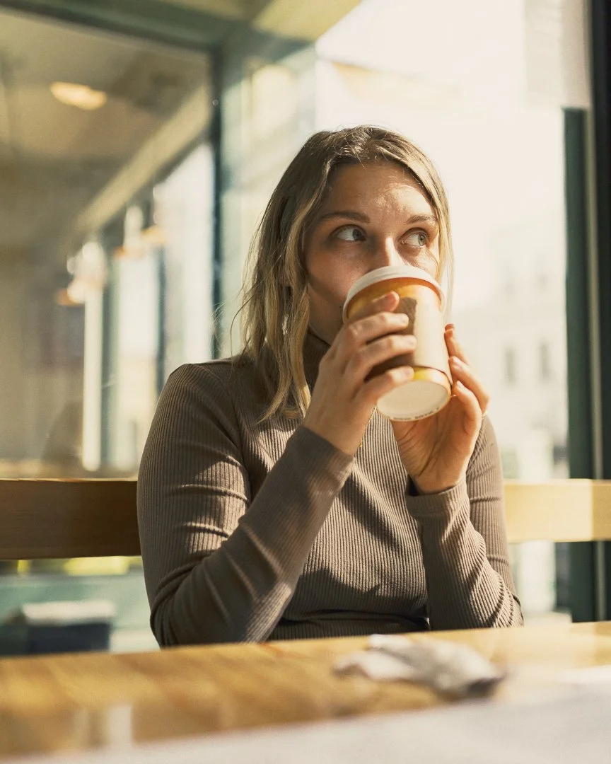A woman sitting in a cafe, drinking coffee from a disposable cup by a window on a sunny day.