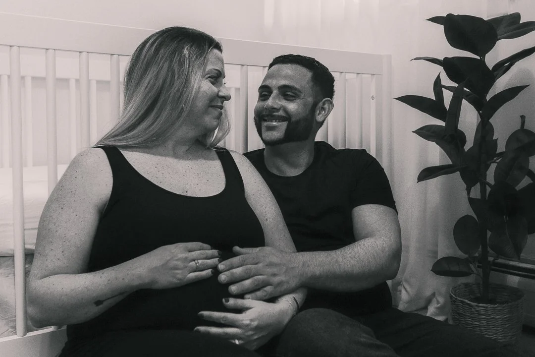 A woman and a man sitting on a bed, looking at each other and smiling. The woman is pregnant, and the man is holding her belly. A potted plant is visible on the right side.