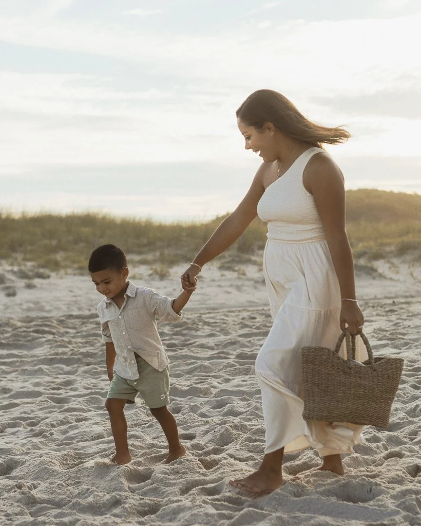 A woman and a young boy holding hands and walking barefoot on a sandy beach with grass in the background, during sunset.