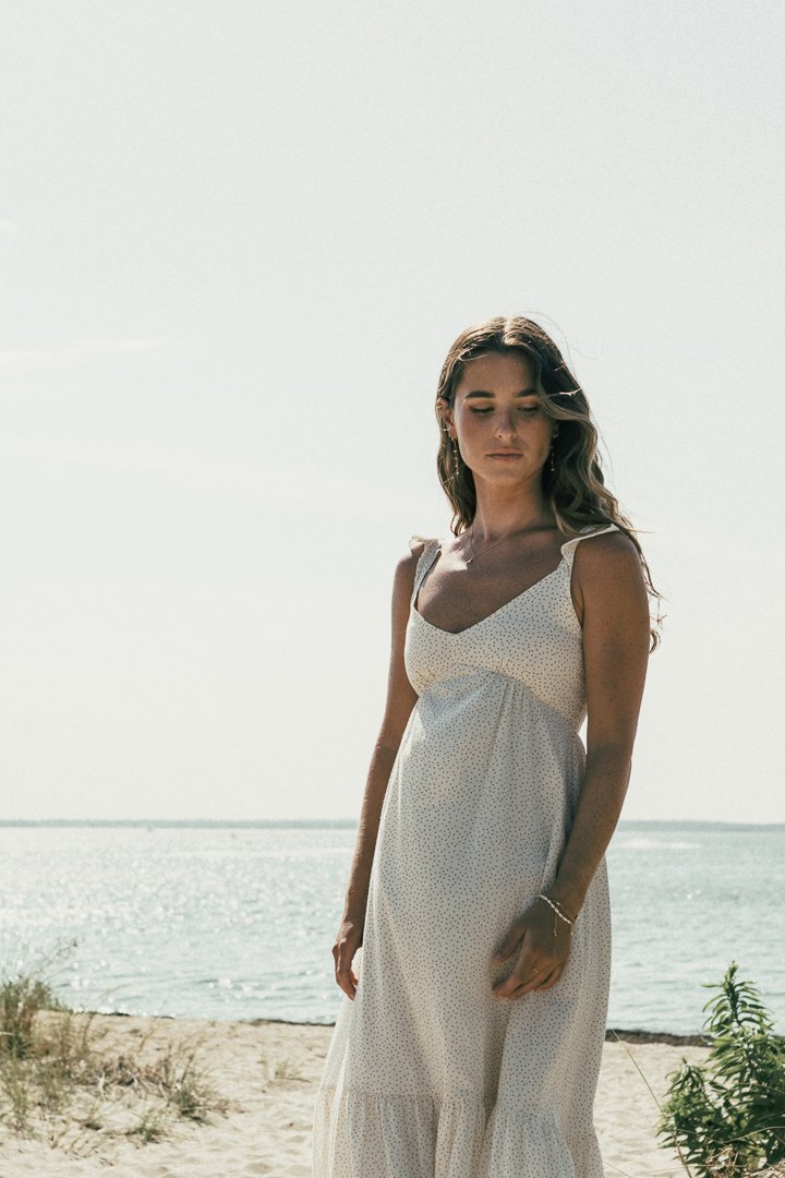 A woman in a white, dotted dress standing on a sandy beach near the water, with the ocean and sky visible in the background.