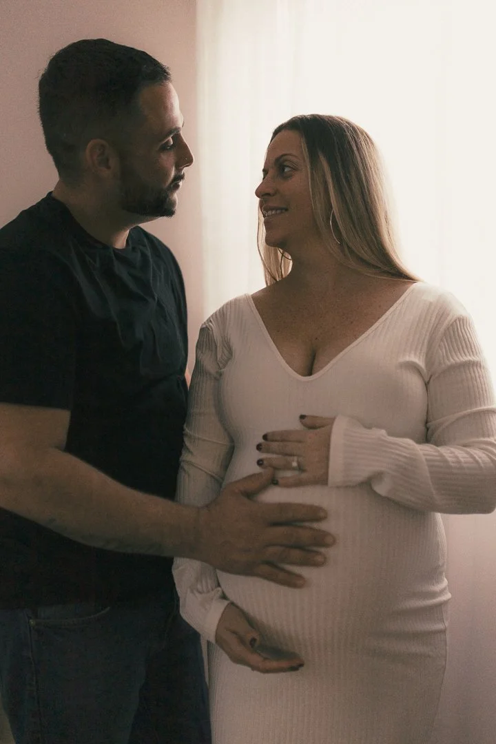 A pregnant woman and a man touching her belly, standing close and looking at each other affectionately indoors.