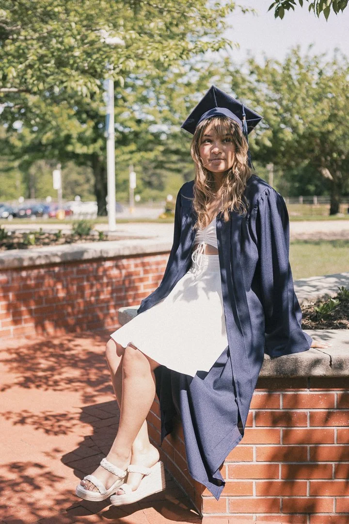 A young girl in a graduation gown and cap sitting on a brick wall outdoors, with trees and a parking lot in the background.